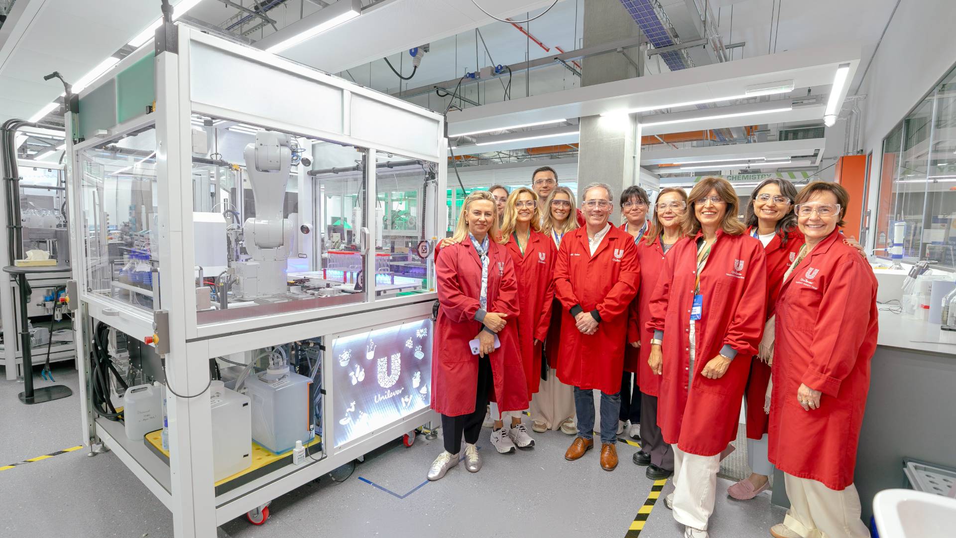 Kırmızı önlük giyen kadın ve erkeklerden oluşan on bir kişi robot laboratuvarında poz veriyor.