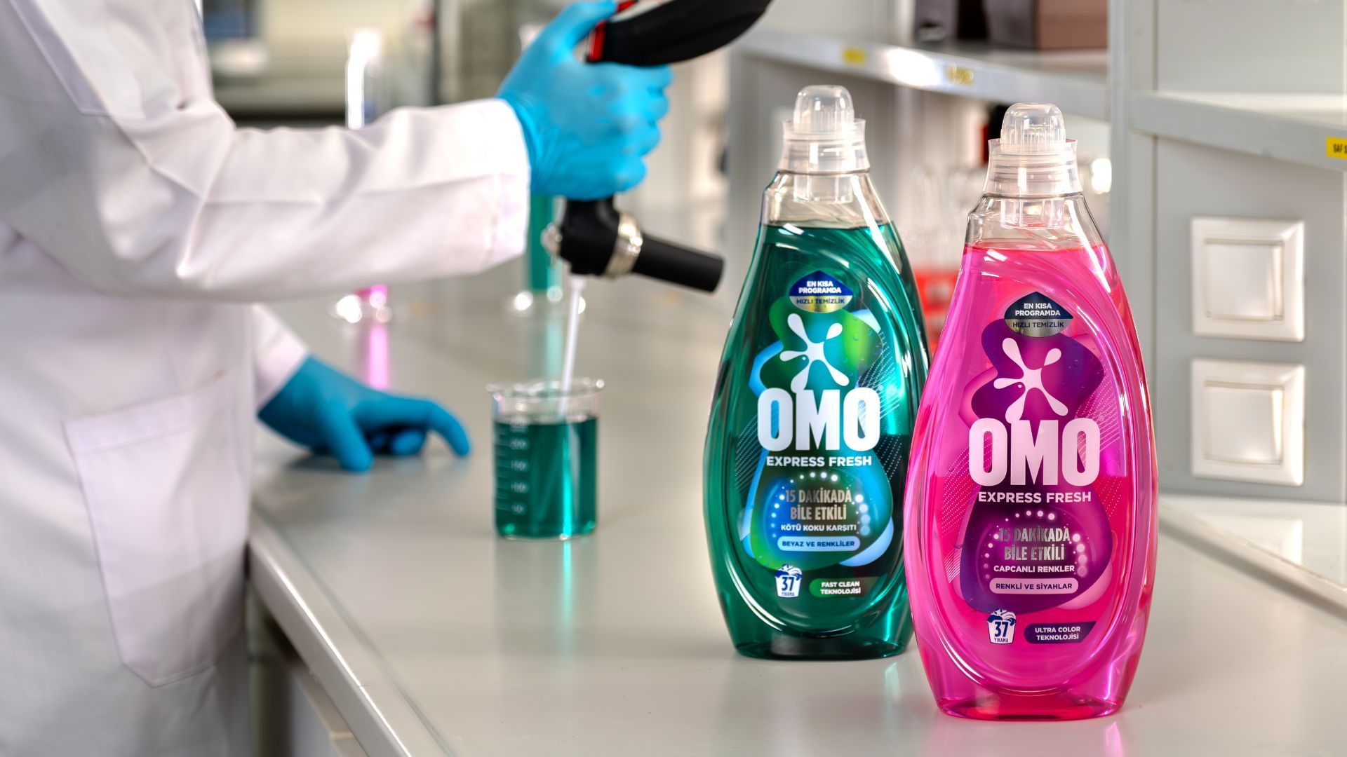 Sağ önde Omo Express Fresh’in yeşil ve pembe iki şişesi var, sol arkada laboratuvar önlüğüyle iki elini gördüğümüz bir araştırmacı ölçüm yapıyor. 